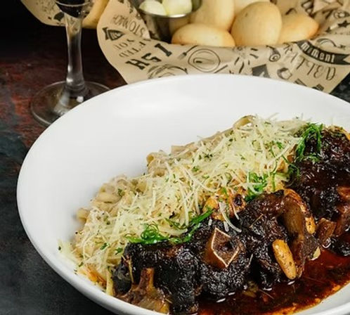 Braised short ribs in a glossy red-wine mushroom sauce atop parmesan-flecked fettuccine, garnished with chopped herbs, with bread rolls and a glass of red wine in the background.