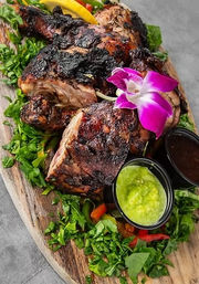 Charred spicy jerk chicken pieces on a wooden serving board with chopped greens, lemon wedge, two dipping sauces (bright green salsa and dark sauce) and a purple orchid garnish — tropical platter.