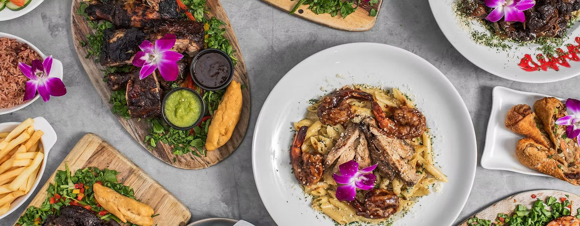 Top-down restaurant table spread of grilled meats and shrimp pasta with sides—fries, rice, salads and fried dumplings—garnished with purple orchids on a gray tabletop.