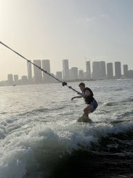 Wakeboarder being towed across choppy water with a high-rise coastal city skyline and soft sunset light in the background.