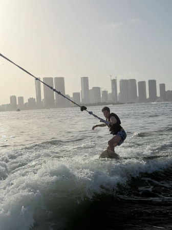 Wakeboarder being towed across choppy water with a high-rise coastal city skyline and soft sunset light in the background.