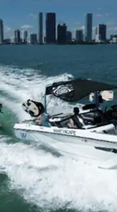 Wakeboarder riding the wake behind a white motorboat speeding across turquoise bay with a modern city skyline on the horizon under a sunny blue sky