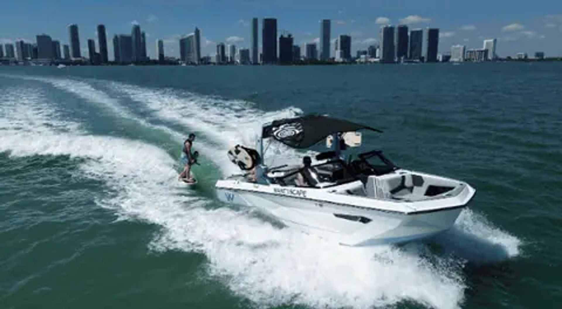 Wakeboarder riding the wake behind a white motorboat speeding across turquoise bay with a modern city skyline on the horizon under a sunny blue sky
