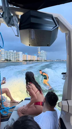 Wakesurfer riding a boat wake at dusk while friends watch from the stern, illuminated coastal high-rise skyline in the background