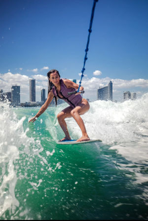 Person wakesurfing on vibrant turquoise water while holding a tow rope, with a coastal city skyline and bright blue sky in the background.