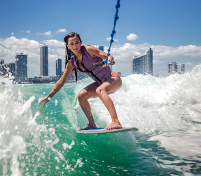 Person wakesurfing on vibrant turquoise water while holding a tow rope, with a coastal city skyline and bright blue sky in the background.