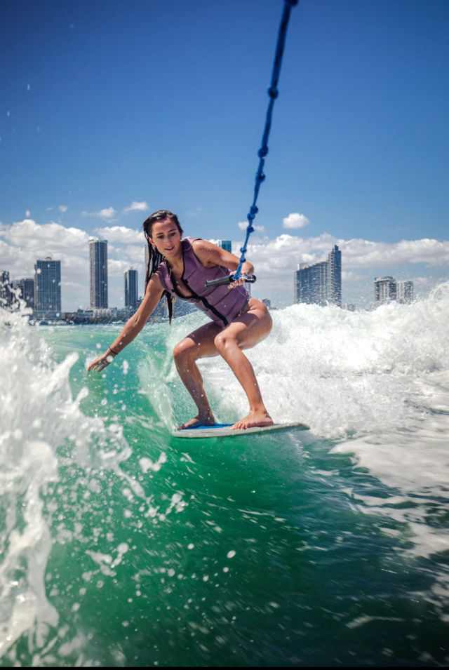 Person wakesurfing on vibrant turquoise water while holding a tow rope, with a coastal city skyline and bright blue sky in the background.