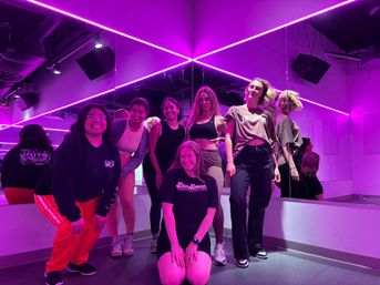 Small group of six people posing in a neon-purple dance studio with mirrored walls, LED strip lights and casual athletic wear — fun group dance class vibe.