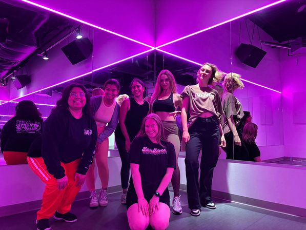 Small group of six people posing in a neon-purple dance studio with mirrored walls, LED strip lights and casual athletic wear — fun group dance class vibe.