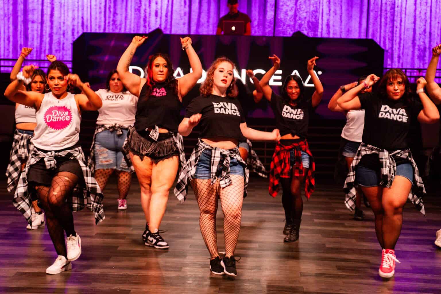Energetic group of dancers performing a synchronized street-style routine on an indoor stage with a purple curtain backdrop, wearing casual streetwear, denim shorts, fishnet tights and plaid shirts tied at the waist.
