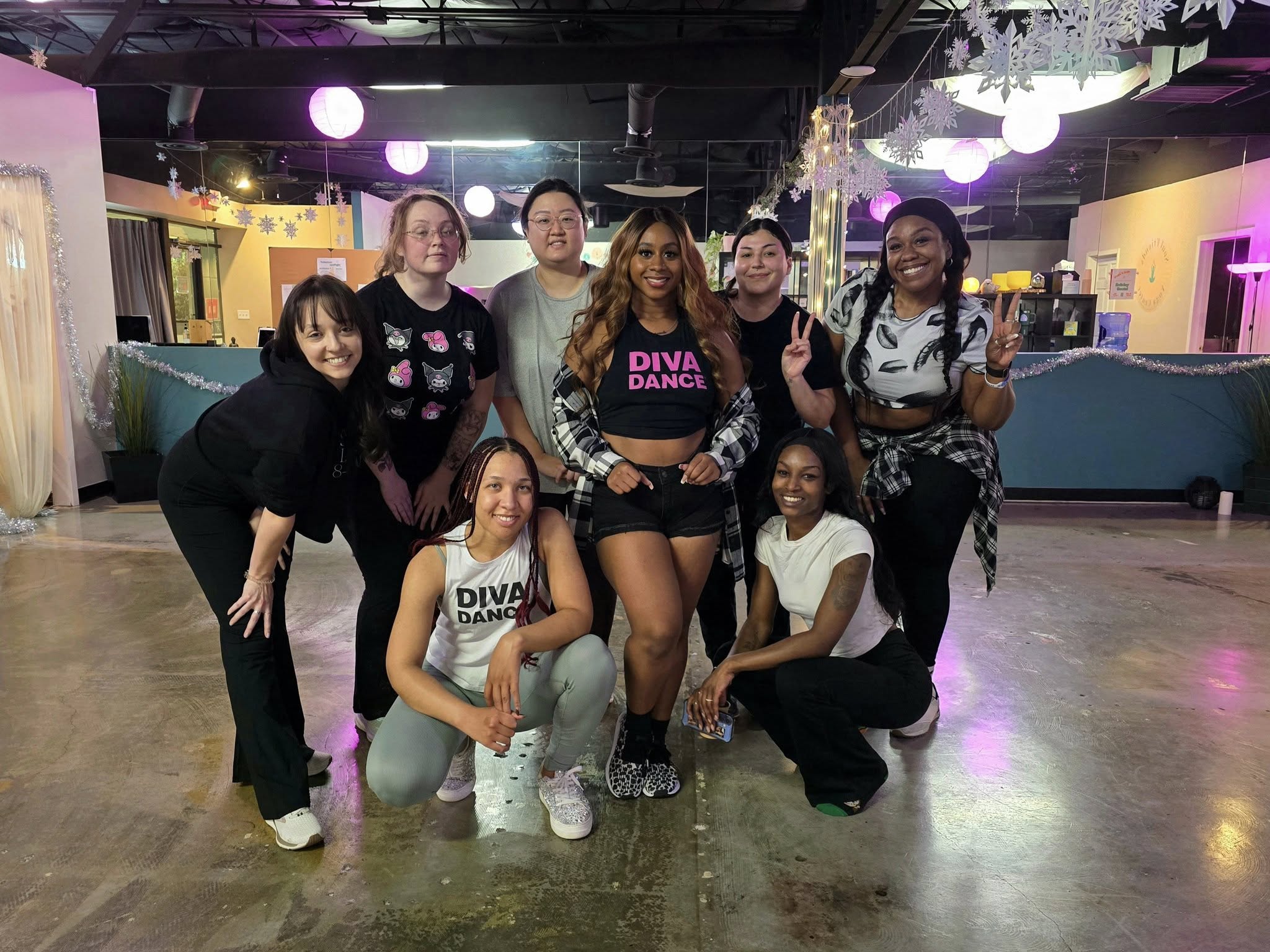 Eight women posing in an indoor dance studio with purple lantern lights and winter decorations, wearing activewear and plaid shirts, smiling and flashing peace signs.