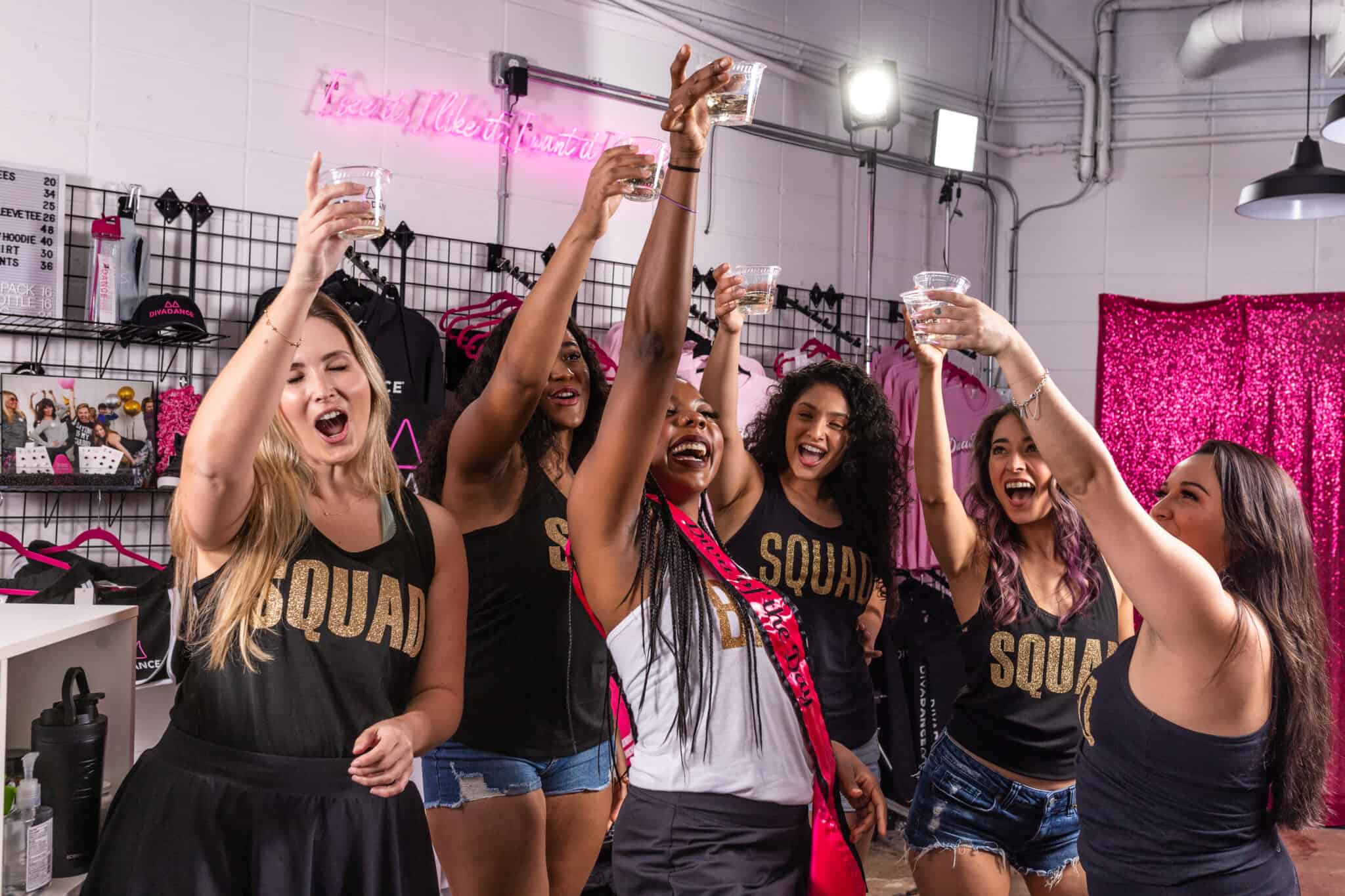 Six women in matching 'SQUAD' tank tops, one wearing a sash, cheer and raise plastic cups for a bachelorette-style toast in a pink-decorated boutique party space with a neon sign and clothing racks.