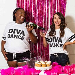 Two smiling dancers in matching white tees and black shorts pour bubbly into plastic cups in front of a pink tinsel backdrop with a cupcake display and bright pink feather decorations.