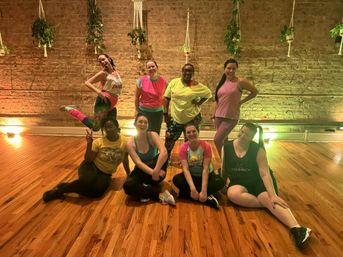 Nine people in colorful workout clothes posing and smiling after a dance class in an exposed-brick, hardwood-floor studio with hanging plants and warm accent lighting.