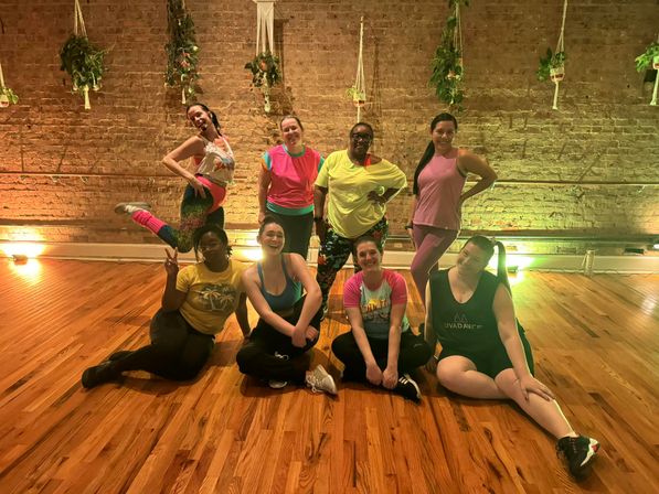 Nine people in colorful workout clothes posing and smiling after a dance class in an exposed-brick, hardwood-floor studio with hanging plants and warm accent lighting.