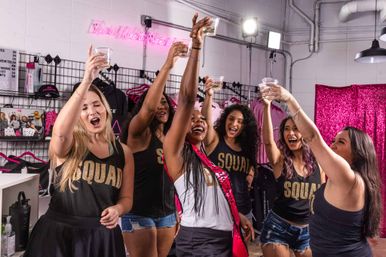 Six friends in matching "SQUAD" tanks raising plastic cups for a bachelorette toast in a pink-decorated party boutique/studio.