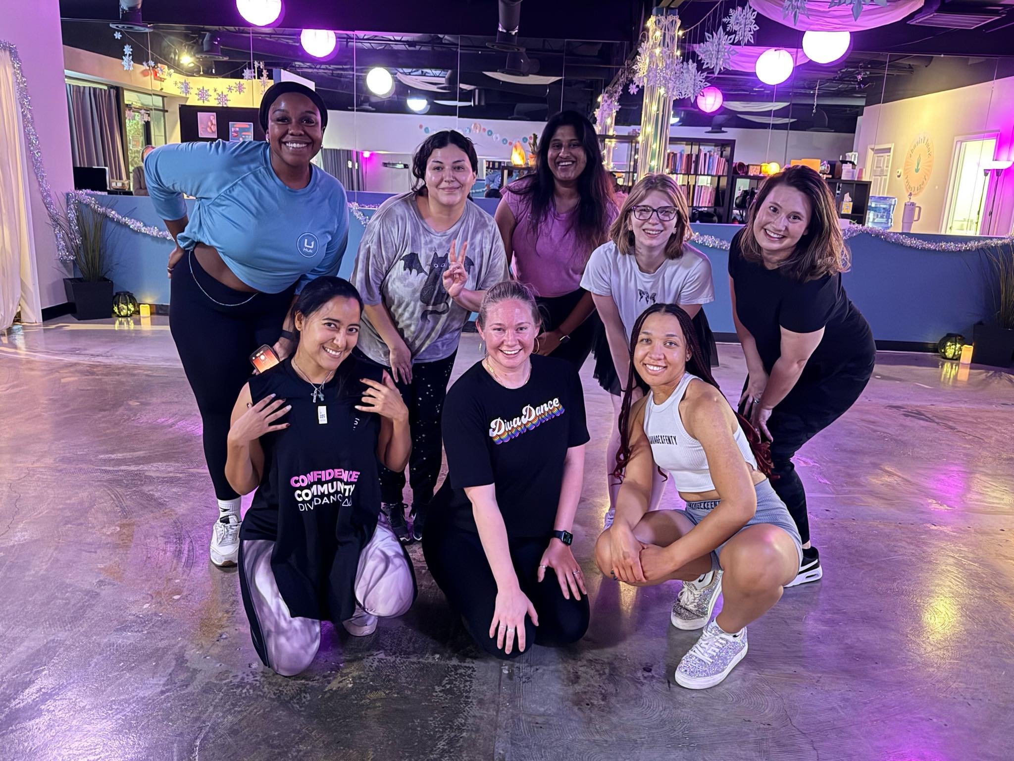 Smiling diverse group of nine women posing in a brightly lit indoor dance studio with purple and blue lighting, mirrored wall, festive decorations, and polished concrete floor — post-dance class group photo.