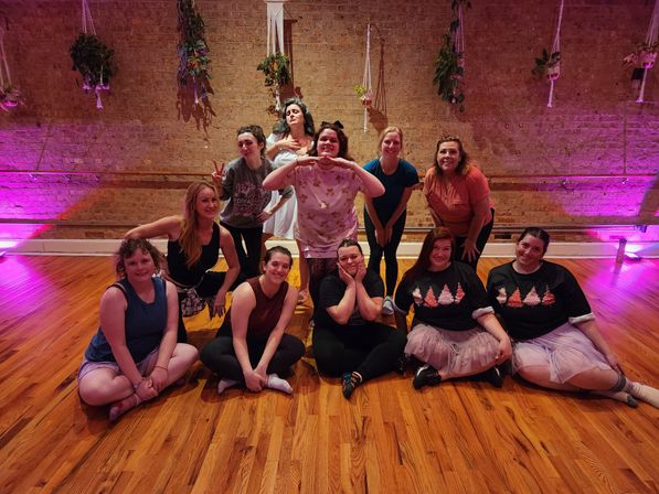 Group photo of people posing in a dance studio with hardwood floors and exposed brick wall, hanging plants and pink-purple uplighting; participants sit and stand in casual workout clothes and tutus, smiling and striking playful poses.