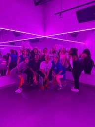 Group workout in a mirrored dance studio lit by neon purple LED lights, smiling participants posing with towels after class.