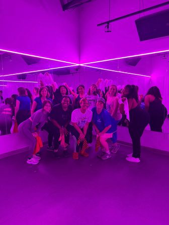 Group workout in a mirrored dance studio lit by neon purple LED lights, smiling participants posing with towels after class.