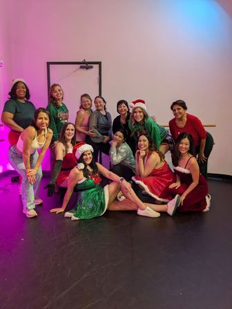 Group of women in holiday costumes and Santa hats posing and smiling in a dance studio by a ballet barre for a festive group photo.