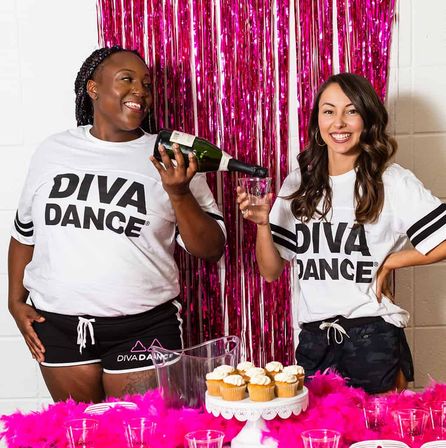 Two smiling women in matching white logo shirts pouring champagne at an indoor party with a pink tinsel backdrop, cupcakes on a stand, clear cups and bright pink feather table decor.