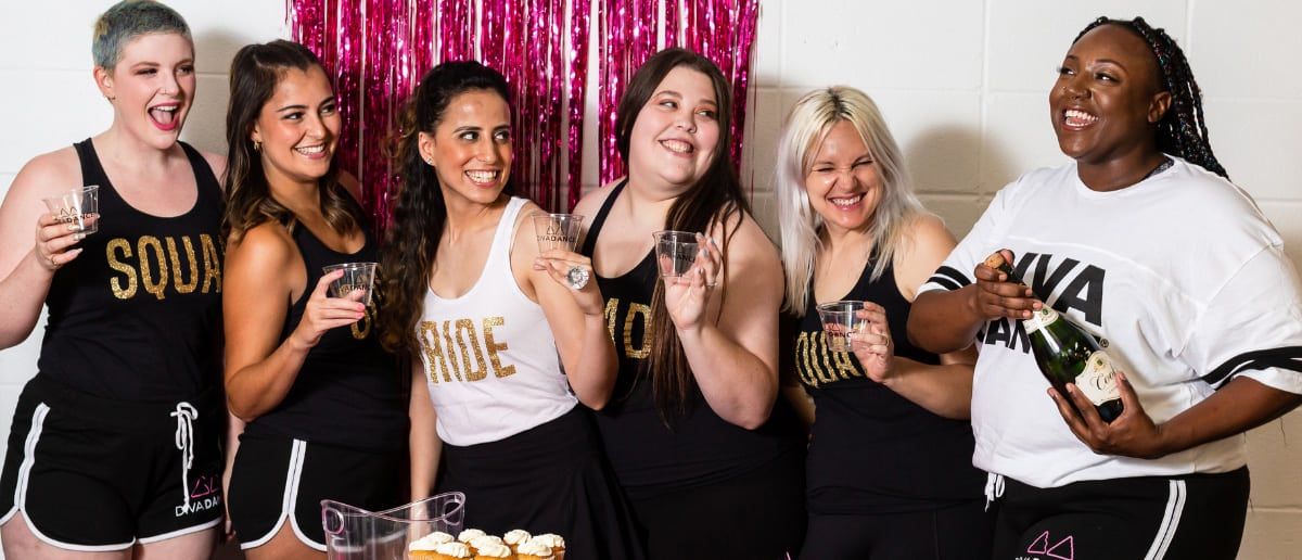 Six friends at a bachelorette party laughing and toasting — bride in a white "BRIDE" tank with friends in matching black "SQUAD" tops, holding drinks and popping champagne in front of a pink fringe backdrop with cupcakes on the table.