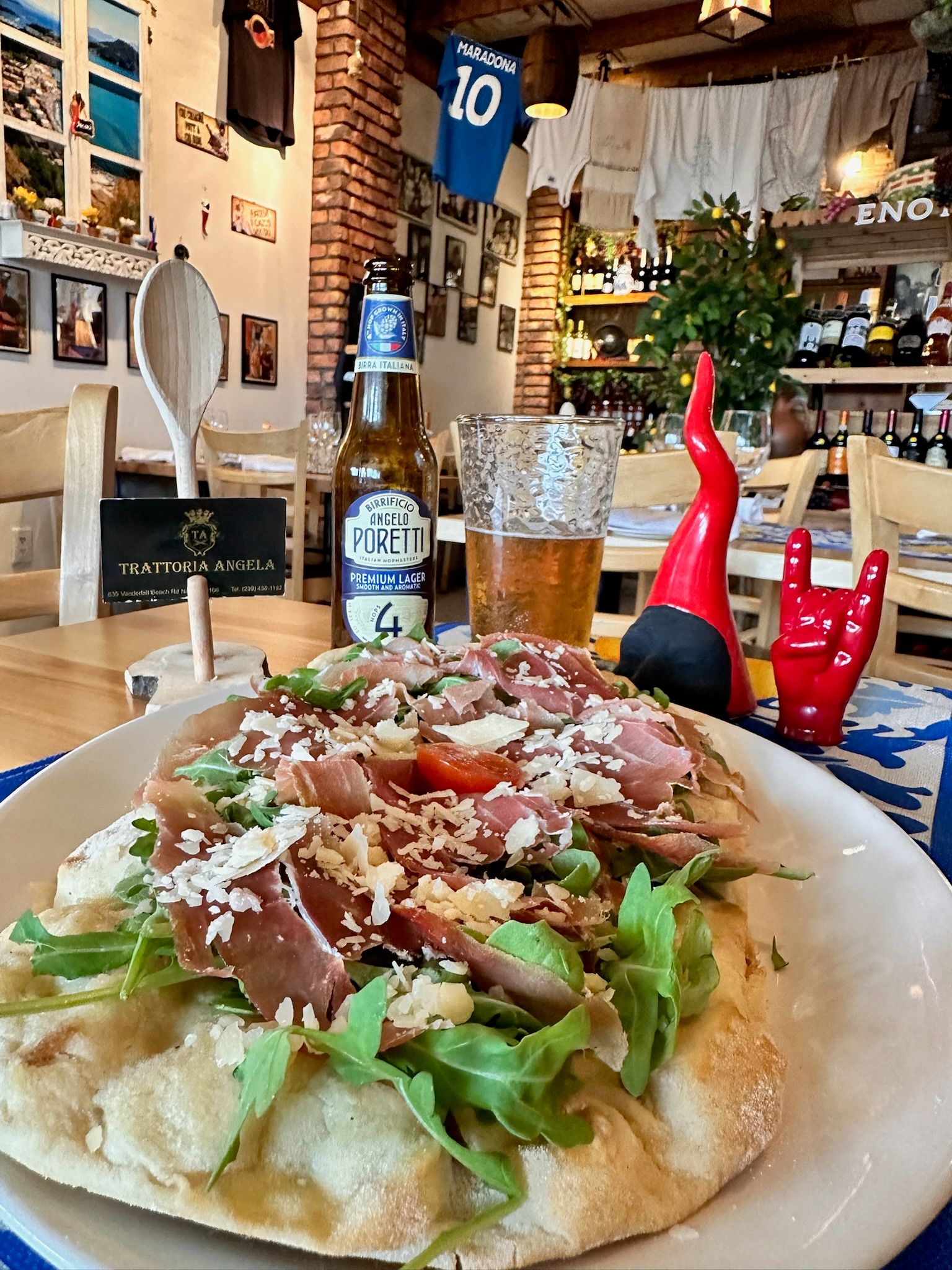 Prosciutto and arugula flatbread topped with shaved Parmesan and a cold lager on a wooden table in a cozy rustic Italian trattoria interior with brick walls and wine bottles.
