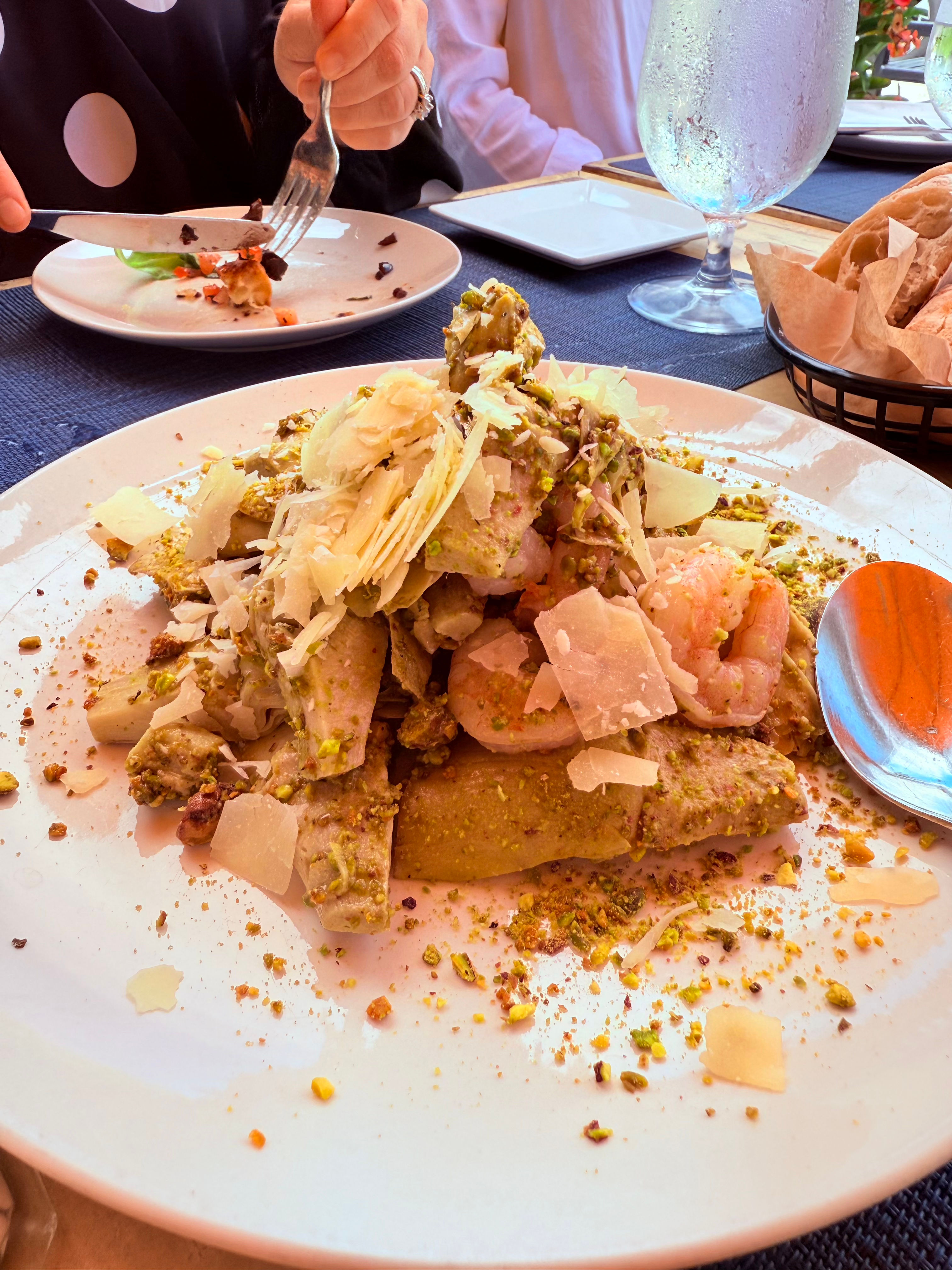 Gourmet shrimp pasta tossed in pistachio pesto, topped with shaved Parmesan and crushed pistachios on a white plate at a restaurant table with bread basket and water glass.