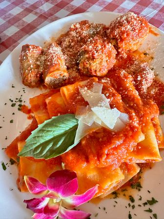 Marinara-smothered Italian ravioli with meatballs, grated Parmesan, basil leaf and orchid garnish on a white plate over a red-checked tablecloth.