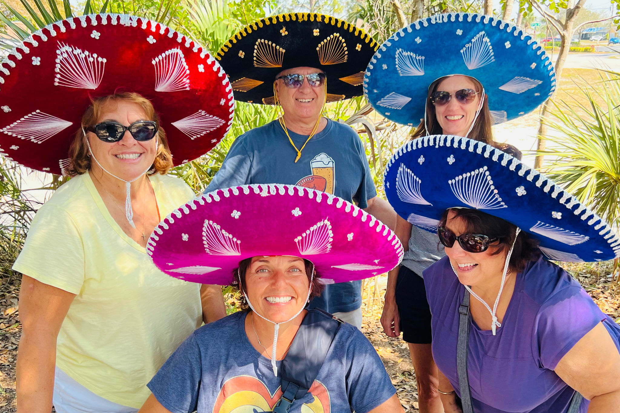 Playful group photo of five adults wearing colorful sombreros (red, pink, black, blue) and sunglasses, smiling outdoors among palm trees on a sunny day.