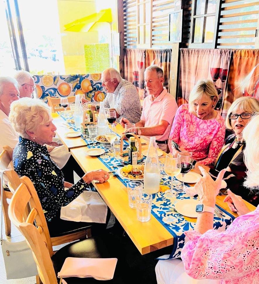 Sunlit restaurant scene of a cheerful group dining at a long wooden table with a blue patterned runner, wine glasses, bottled water, small plates and a colorful citrus mural.
