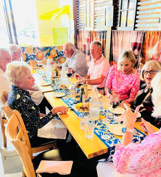 Sunlit restaurant scene of a cheerful group dining at a long wooden table with a blue patterned runner, wine glasses, bottled water, small plates and a colorful citrus mural.