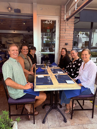 Six people smiling at an outdoor restaurant patio table set with blue placemats and napkins, empty wine glasses and an urban storefront in the background.