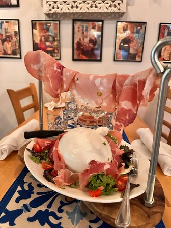 Creamy burrata ball on mixed greens and cherry tomatoes, surrounded by prosciutto slices suspended from a metal rack as a playful Italian antipasto on a wooden restaurant table with wine glasses and framed photos in the background.