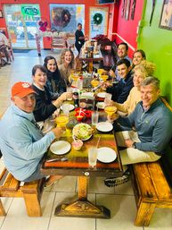 Group of friends laughing and toasting margaritas at a long wooden table in a colorful casual Mexican restaurant with red and green walls and holiday wreaths.