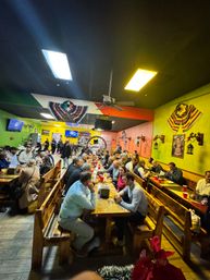 Lively, busy Mexican restaurant interior with colorful green and yellow walls, long communal wooden benches and tables packed with diners, festive serapes and wall art, ceiling fan and warm lighting creating a bustling group dining scene.