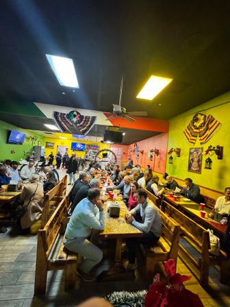 Lively, busy Mexican restaurant interior with colorful green and yellow walls, long communal wooden benches and tables packed with diners, festive serapes and wall art, ceiling fan and warm lighting creating a bustling group dining scene.