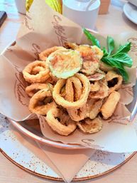 Crispy fried calamari rings and battered zucchini chips garnished with fresh basil, served in a cafe-style bowl on parchment as a golden appetizer on a rustic plate.