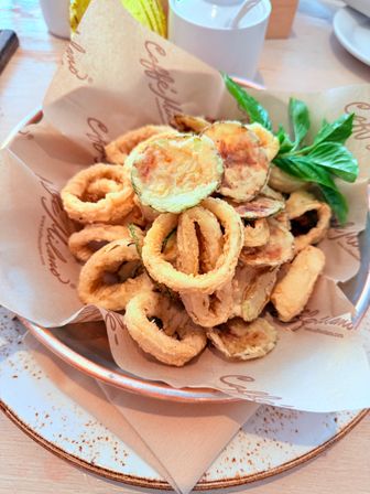 Crispy fried calamari rings and battered zucchini chips garnished with fresh basil, served in a cafe-style bowl on parchment as a golden appetizer on a rustic plate.