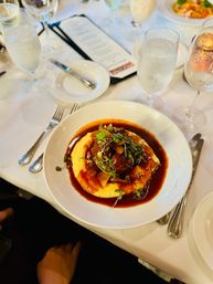 Braised short ribs in a rich red-wine jus served over creamy mashed potatoes, topped with microgreens on a white-tablecloth restaurant table with water and wine glasses and a menu nearby.
