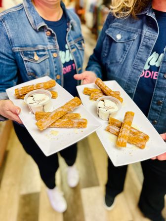 Two people in denim jackets holding plates of crispy golden cinnamon‑sugar churros with vanilla dipping cream and chocolate drizzle at an indoor food market