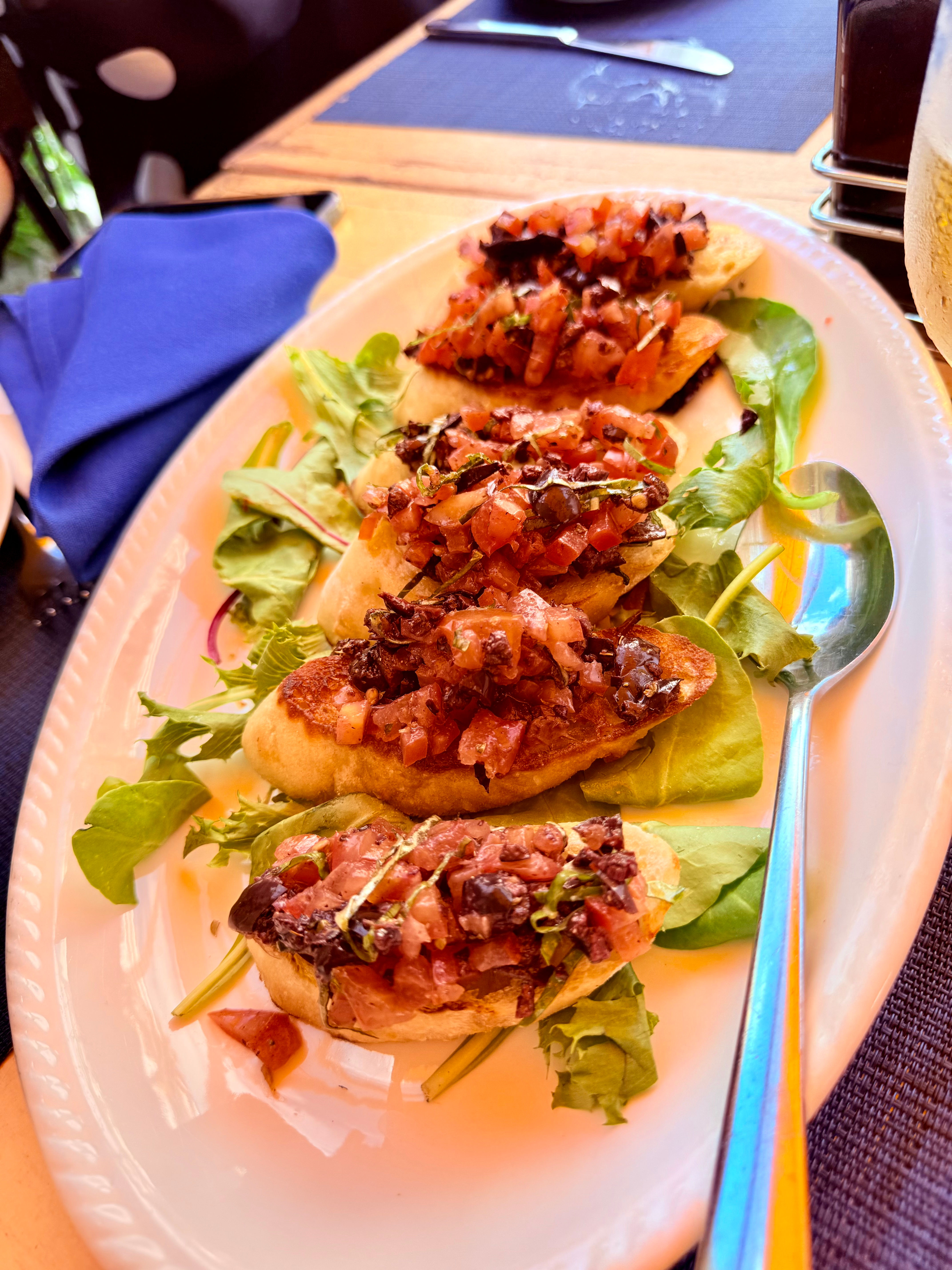 Crispy tomato and basil bruschetta crostini on toasted baguette slices over mixed greens on a white platter, served on a restaurant table with a blue napkin.