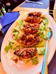 Crispy tomato and basil bruschetta crostini on toasted baguette slices over mixed greens on a white platter, served on a restaurant table with a blue napkin.
