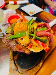 Sizzling cast-iron fajita skillet with grilled steak, chicken, red and green bell peppers, charred scallions and melted cheese on a wooden serving board at a restaurant table.