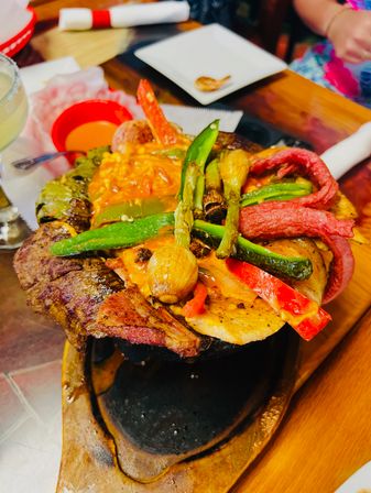 Sizzling cast-iron fajita skillet with grilled steak, chicken, red and green bell peppers, charred scallions and melted cheese on a wooden serving board at a restaurant table.