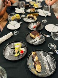 Assorted plated desserts on a black-tablecloth restaurant table — slices of cake, chocolate-filled cannoli, whipped cream with cherries, powdered sugar dusting, wine glasses and rolled napkins.