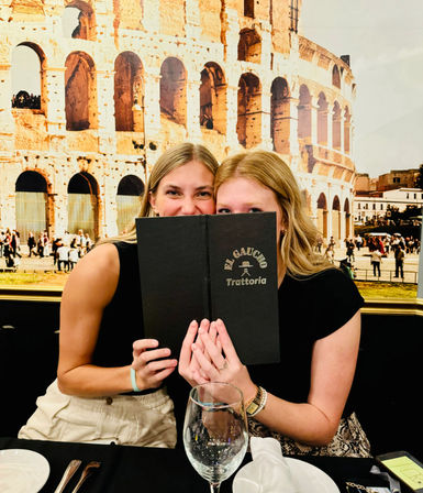Two women playfully peeking over a black menu at an Italian trattoria table, Colosseum mural backdrop, empty wine glass and place settings in the foreground.