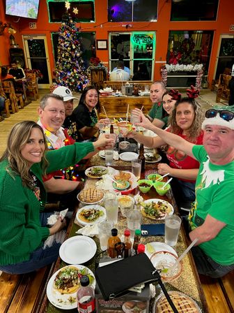 Eight friends toasting over tacos and margaritas at a festive, colorful casual Mexican restaurant interior with a decorated Christmas tree, holiday sweaters and twinkling lights.