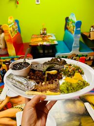 Colorful Mexican restaurant interior with bright green wall and painted booths, plate of Mexican food featuring grilled steak, seasoned crumbled meat, black beans, foil-wrapped tortilla, guacamole with chips and chopped vegetable salsa.